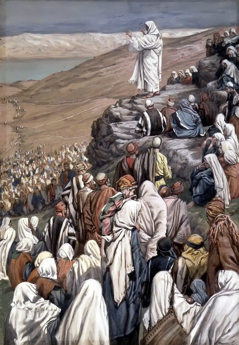 Jesus stands in white robes on a rock high on a hillside with His hands raised as He speaks, while a great crowd in robes and head coverings sits and stands on the slope below Him, with a lake and pale hills behind.