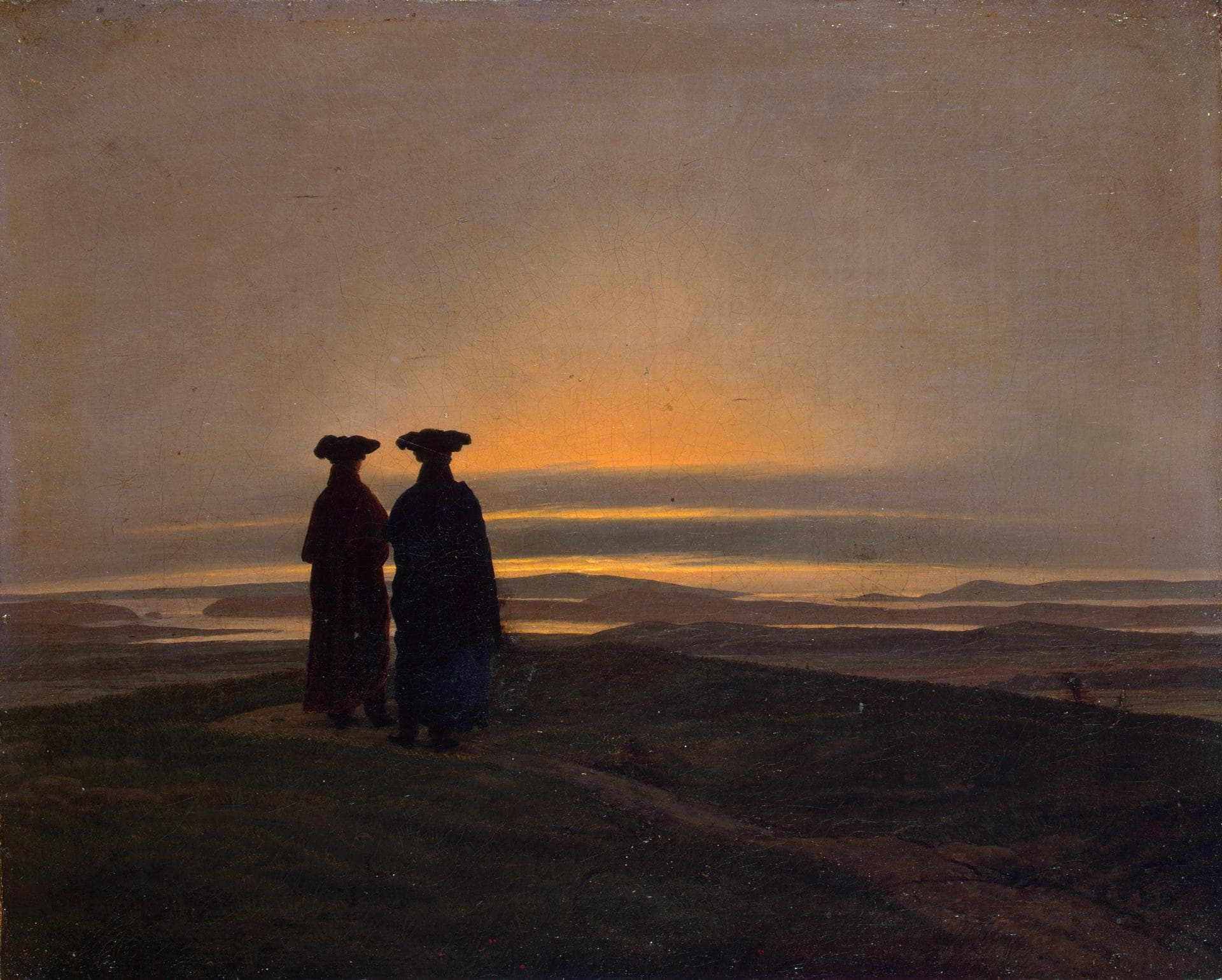 Two men in long dark coats and wide hats stand on a hilltop with their backs to the viewer, watching the sun go down over a wide landscape of water and low hills.