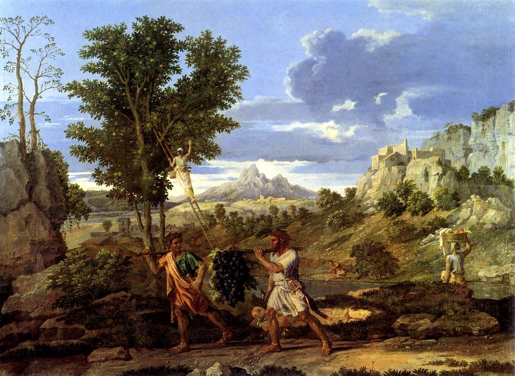 Two men in short tunics carry a huge cluster of dark grapes slung from a pole across a rocky valley, while a man on a ladder picks fruit in a tree at the left and a woman carries a basket of fruit on her head toward a hill town under a blue sky with grey clouds
