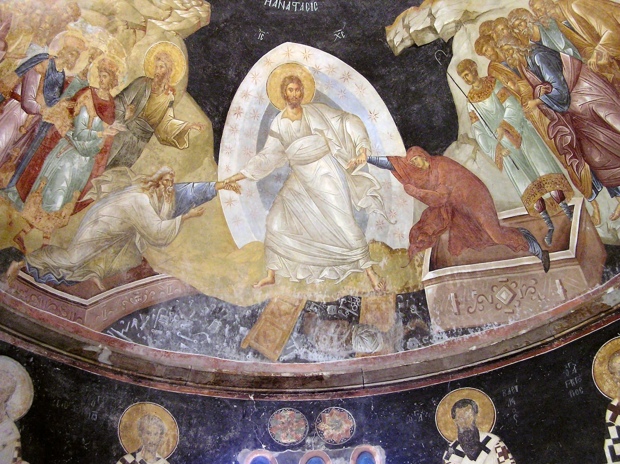 A fresco of Christ in white robes within a starry oval of light, pulling an old man and a woman in red up out of open tombs by the wrist, while broken doors, locks and keys lie scattered on the dark ground beneath His feet and haloed figures crowd on either side.