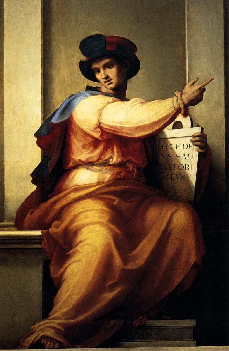 The prophet Isaiah, young and seated in an orange robe with a blue cloak and a dark hat, turns his head and points upward with his right hand while his left holds a tablet inscribed in Latin, before a grey stone niche.