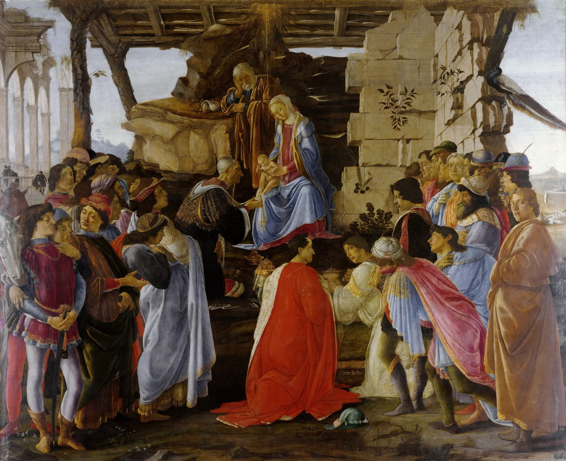 Men in rich robes and hats of many fashions crowd around a ruined stone shelter, where Mary sits holding the infant Jesus and Joseph stands behind her. An old man kneels at the child's feet, and two more kneel in the foreground, one in a red cloak.