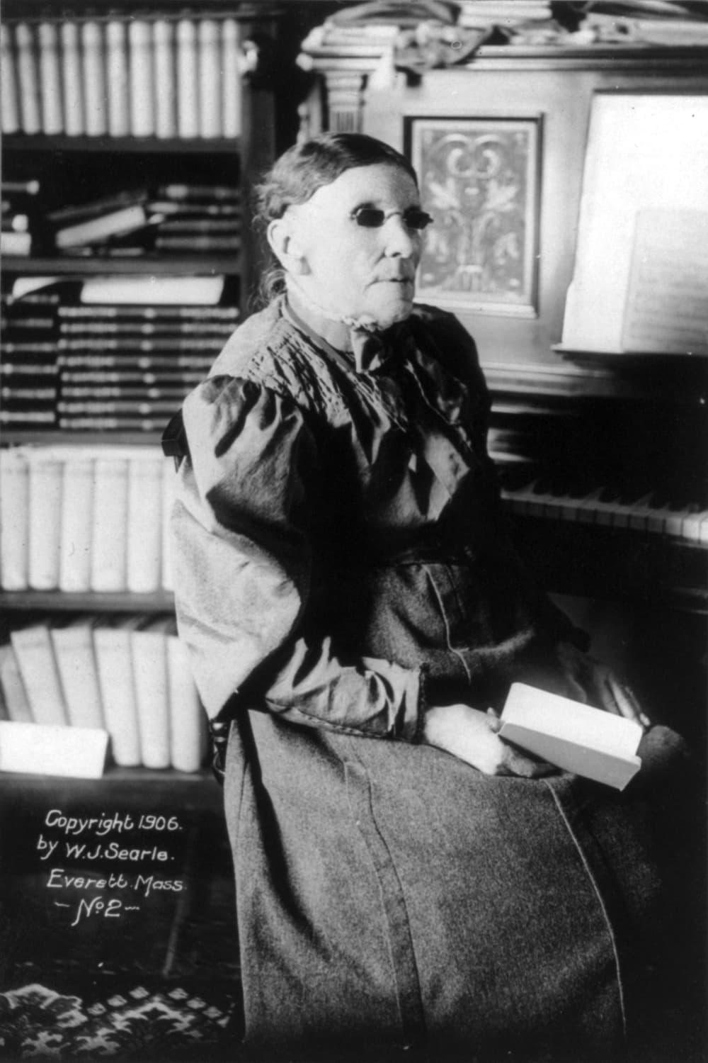 A black and white photograph shows an elderly woman in dark spectacles and a dark dress seated beside a piano, with a book in her hands and shelves of books behind her.