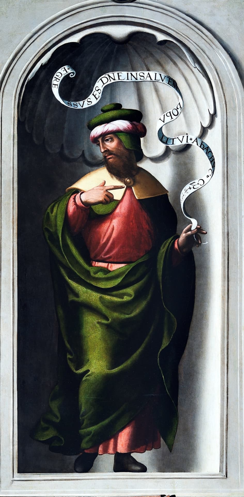 A bearded prophet in a green and rose turban, a red robe and a long green cloak stands in a grey stone niche with a shell carved above him, pointing to his chest with one hand while a Latin scroll bearing his name curls up from the other hand and over his head.