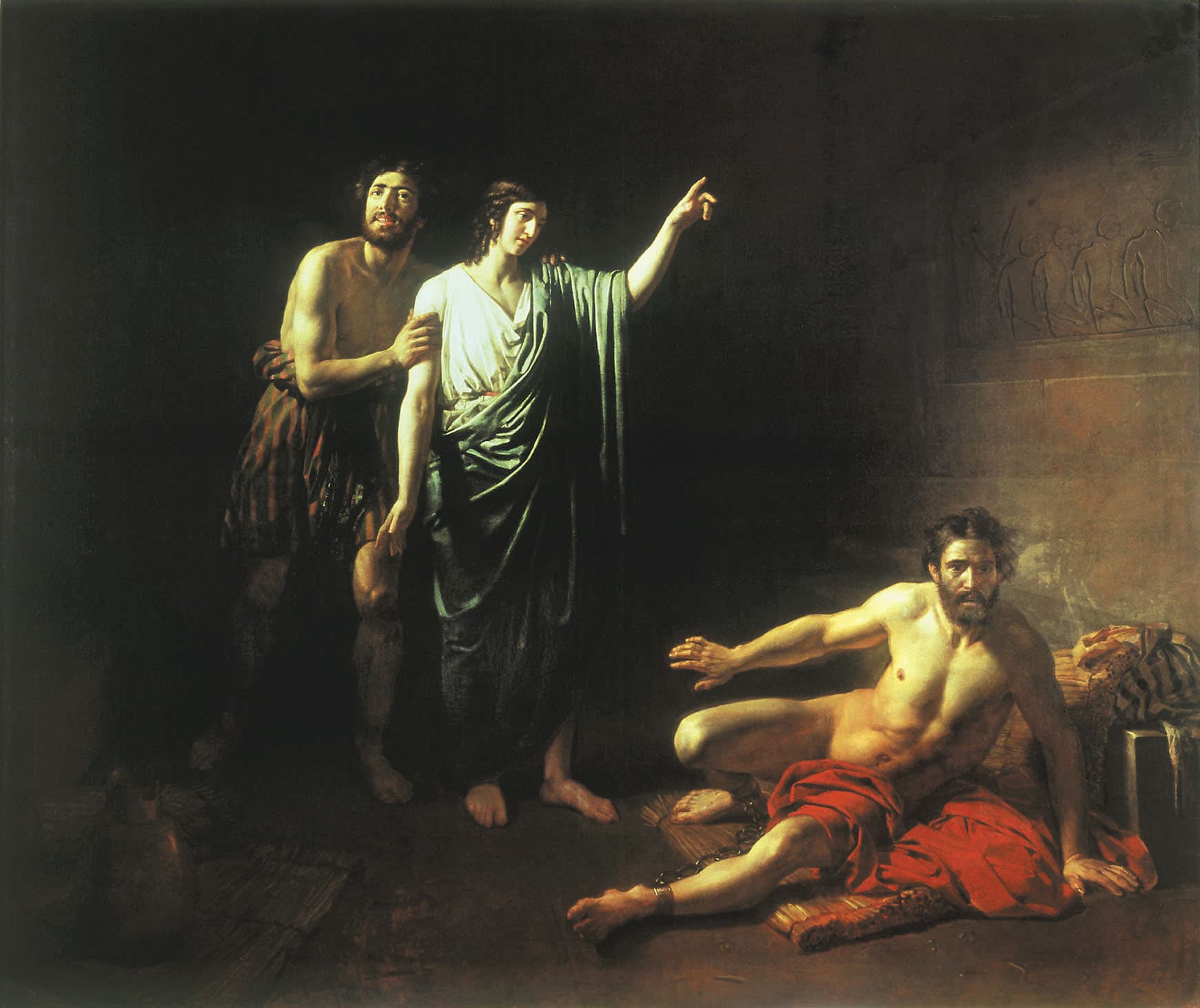 In a dark stone cell, Joseph in a white tunic and green mantle points upward while a smiling bearded prisoner holds his arm, and a second prisoner, chained at the ankle and wrapped in red cloth, sits on straw and throws up his hand in alarm beside a wall carved with figures.