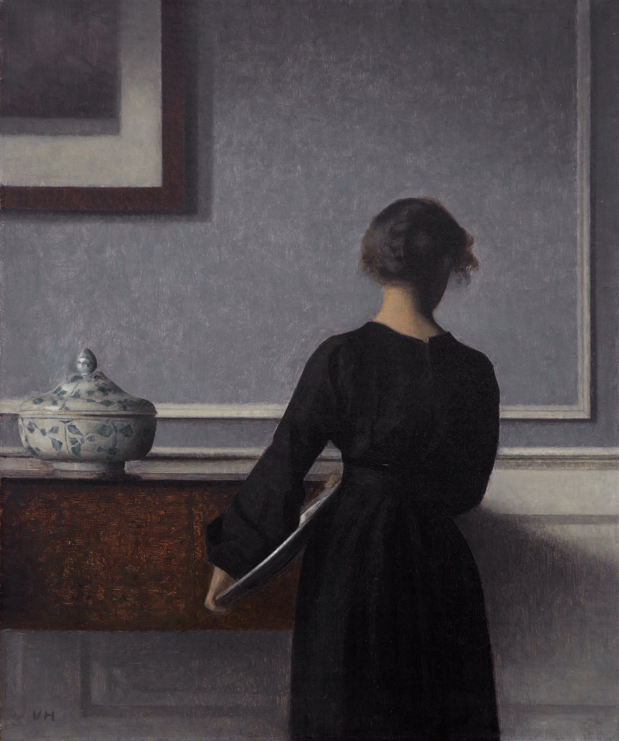 A young woman in a dark dress stands with her back turned in a bare grey room, beside a tall cabinet and a large white bowl on a table.