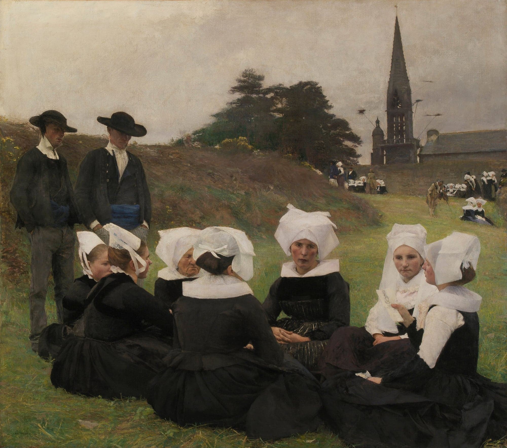 Women in black dresses, wide white collars and tall white linen caps sit in a circle on the grass, and one of them holds a small printed paper. Two men in black jackets, blue sashes and wide black hats stand behind them, and more villagers sit on the grass near a church with a tall stone spire.