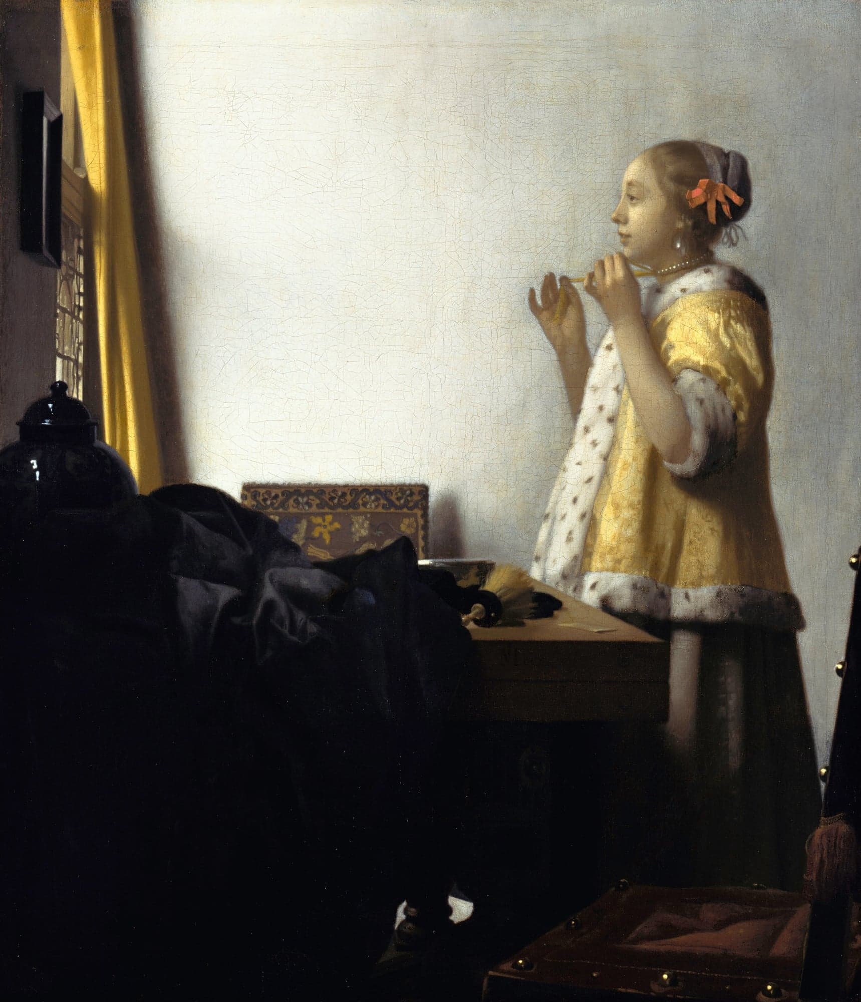 A young woman in a yellow jacket trimmed with ermine stands at a table by a curtained window and holds up the ribbons of a pearl necklace at her throat, facing a small mirror on the wall.
