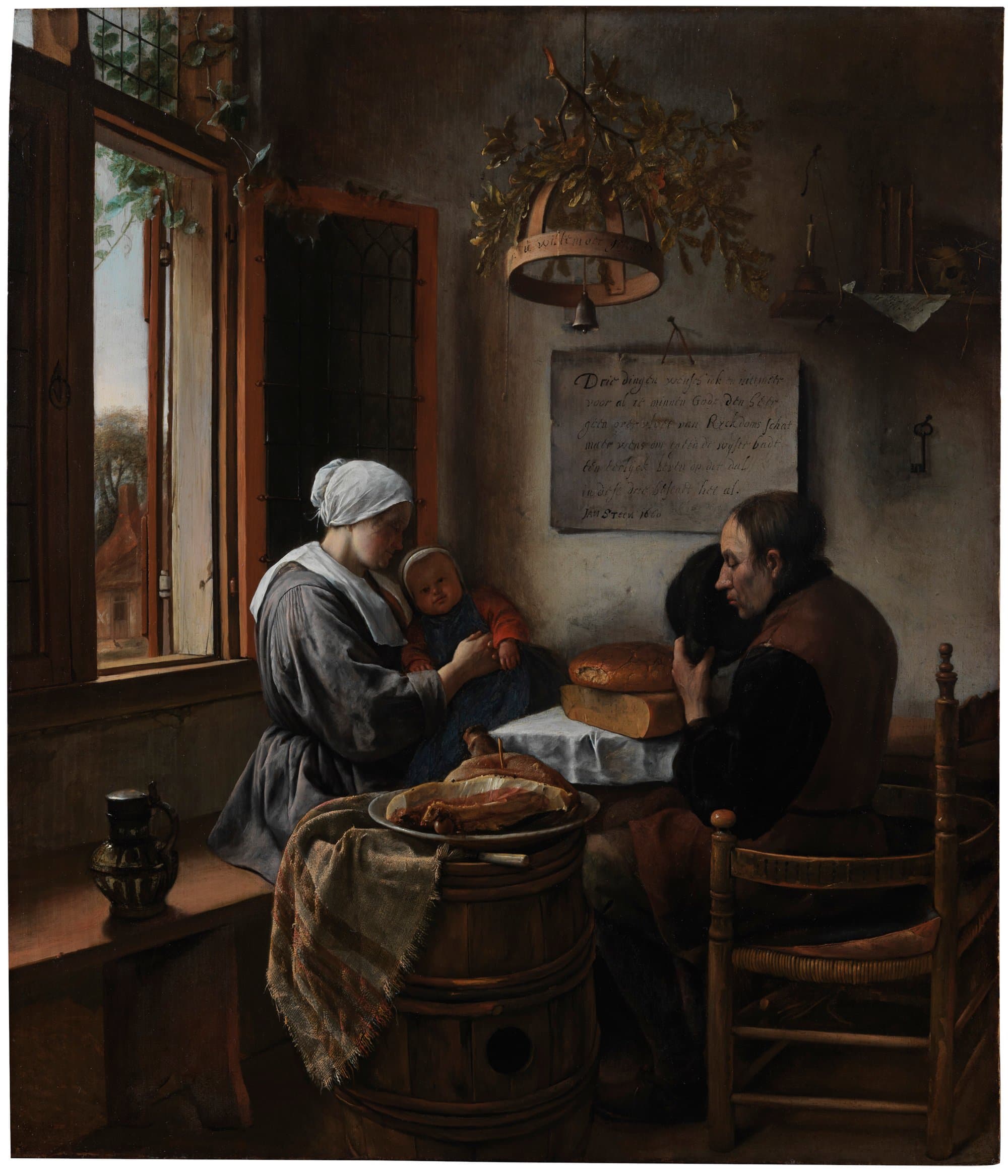 In a plain room lit by an open window, a young mother holding a small child bows her head at a table laid with a white cloth, a loaf of bread and a wedge of cheese, while a man seated on a rush chair holds his hat to his chest and bows his head to pray. A ham lies on a plate on a barrel in front, a stoneware jug stands on the window bench, and a board of written lines hangs on the wall beneath a garland.