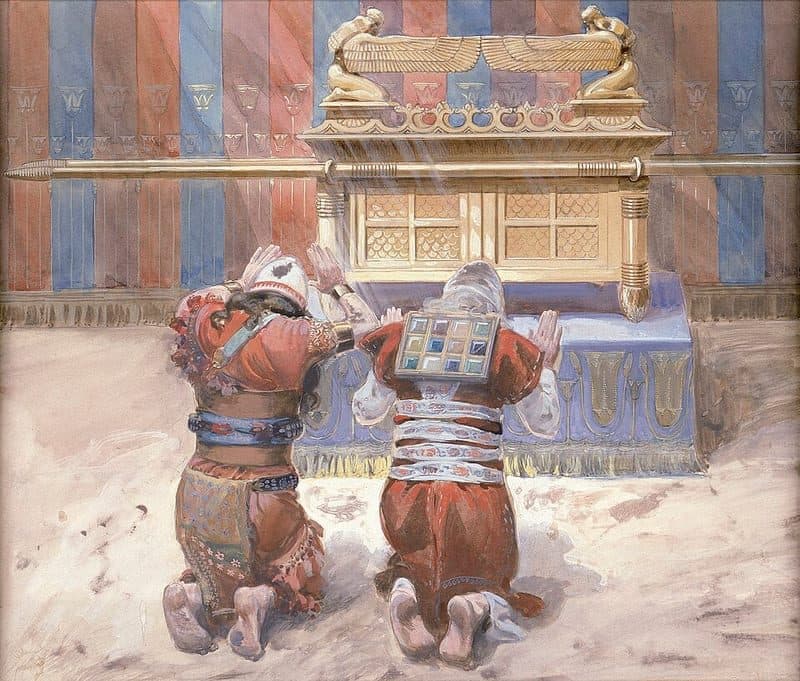 Two men in red robes kneel side by side with their backs to the viewer and their hands raised before a golden ark, whose lid carries two golden winged cherubim, set on a blue cloth inside a tent hung with red and blue striped curtains; one man wears a square panel of twelve coloured stones on his back.