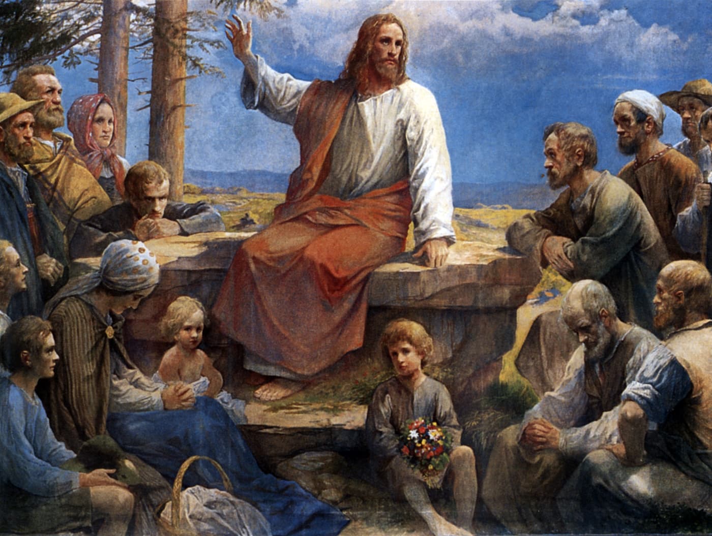 Jesus in a white robe and a red mantle sits on a rock on a hillside with one hand raised, teaching a crowd of men, women and children in country clothes who sit and stand around Him, a boy at His feet holding a bunch of flowers.