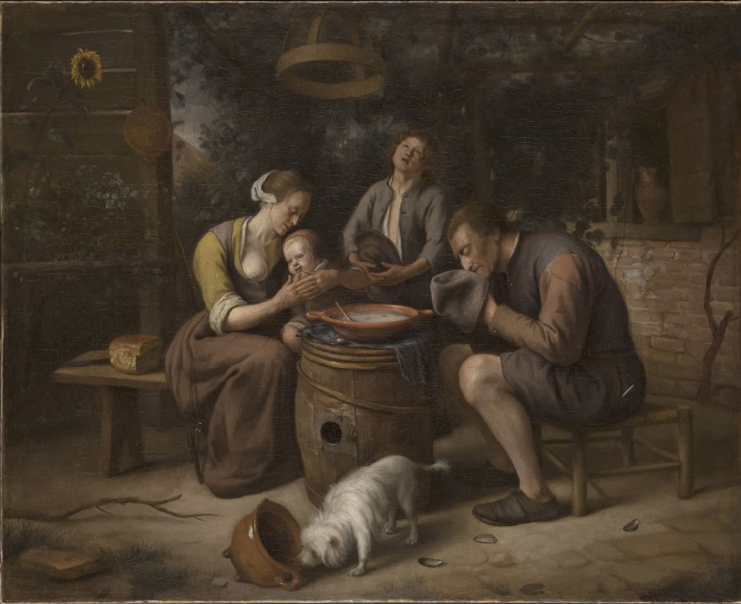 In a farmyard a father bows his head into his hat, a boy looks up, and a mother holds a small child's hands together while a white dog noses an overturned pot.