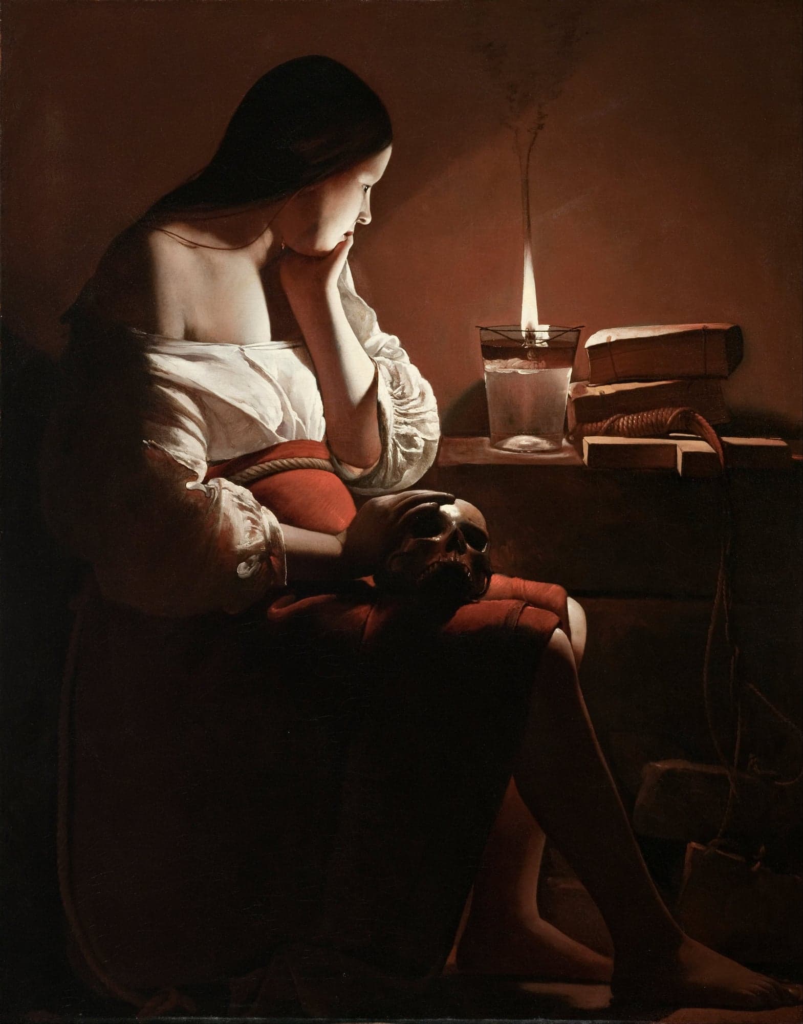A young woman in a white blouse and red skirt sits in a dark room with her chin on her hand, gazing at a candle flame in a glass, a skull resting on her lap and books on the table.