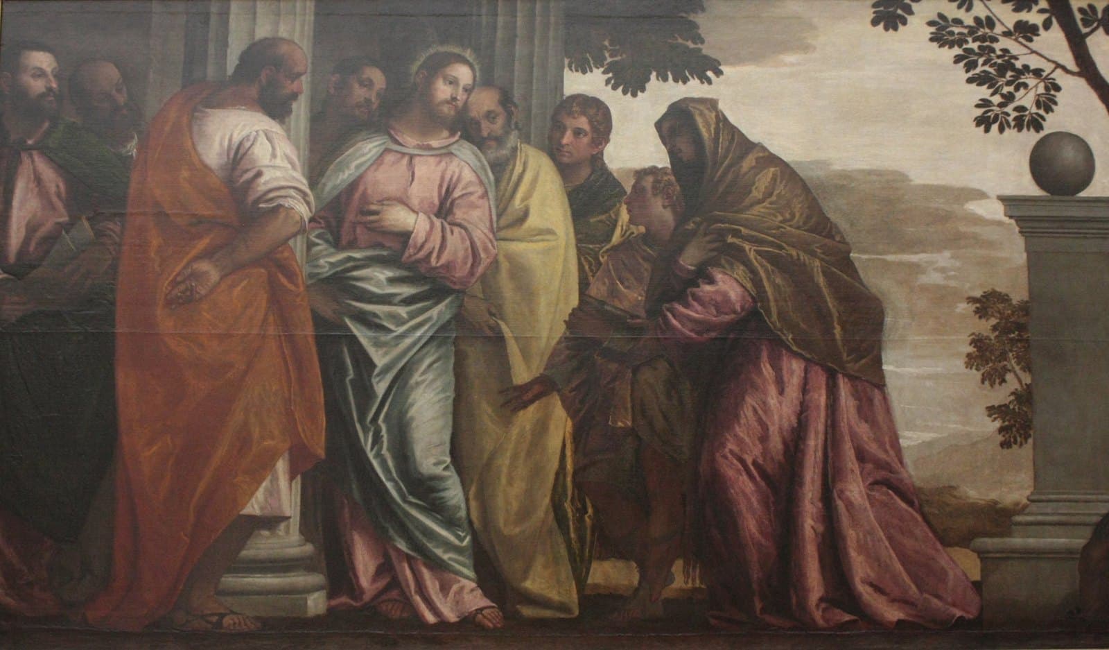 A woman in a pink gown and a gold mantle bends forward with her hand reaching toward Jesus, who stands in a pink robe and a blue-green cloak among His disciples beside a stone column, while a young man at her side looks up at Him and trees and a cloudy sky open behind them.