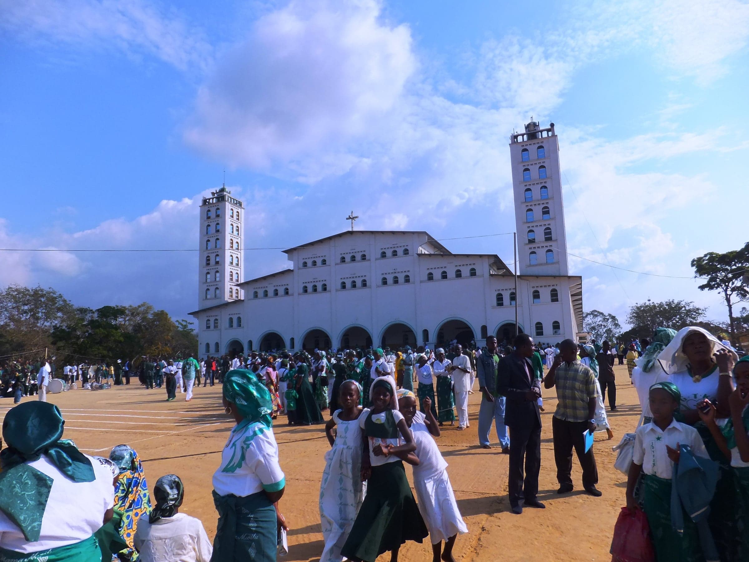 A long white church with two tall square towers stands across a dusty open ground, where people in green and white walk under a bright sky.
