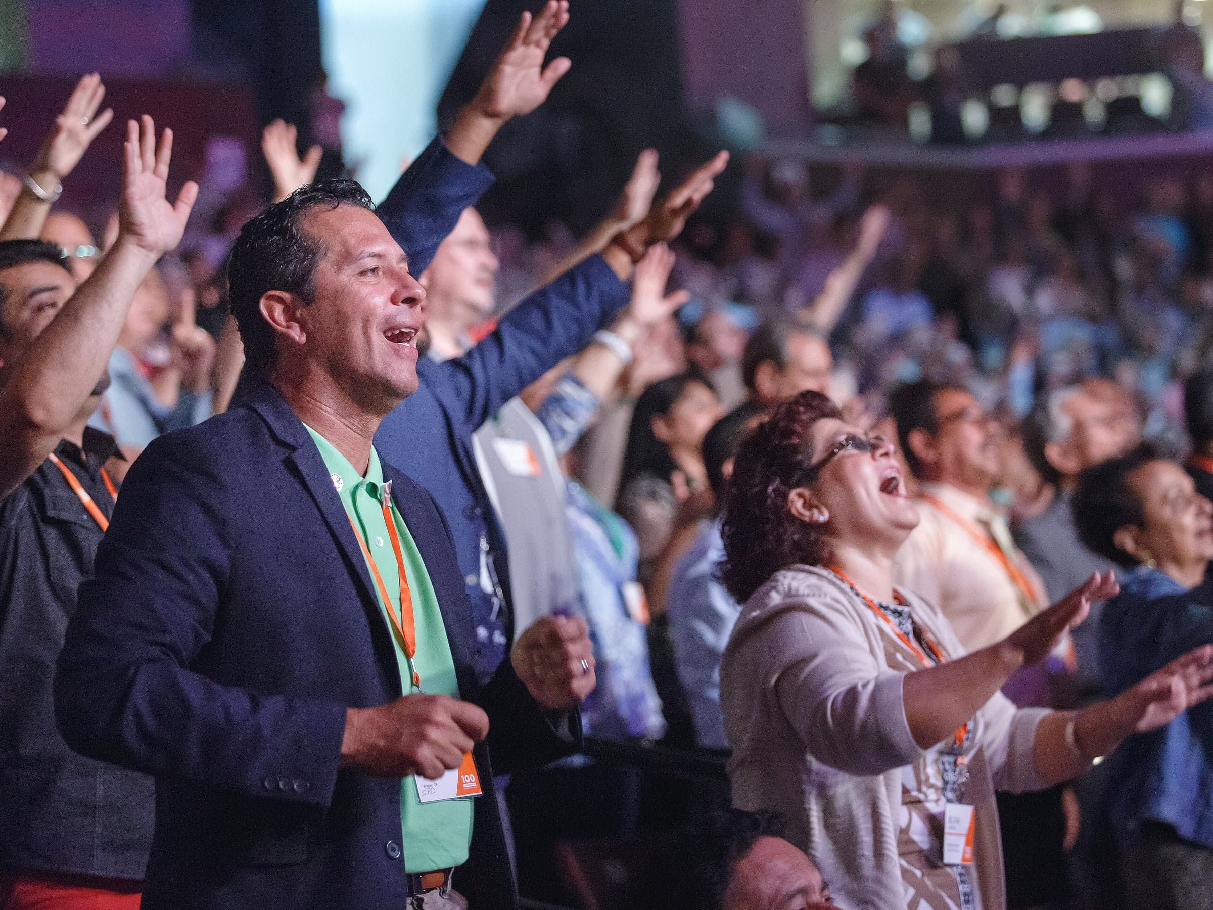 A crowd stands in worship at a large hall, many with hands raised, a man in a dark jacket and a woman singing in the front rows.