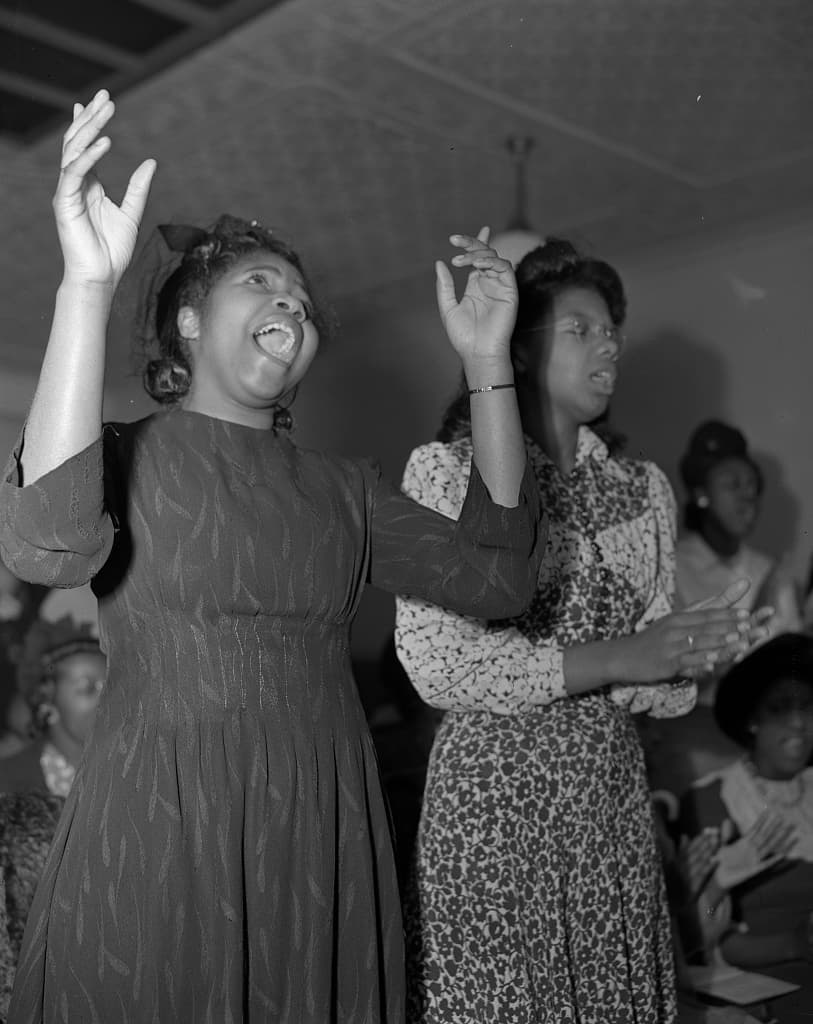 Two women of a Church of God in Christ congregation stand singing, one with both hands raised, others seated behind them.