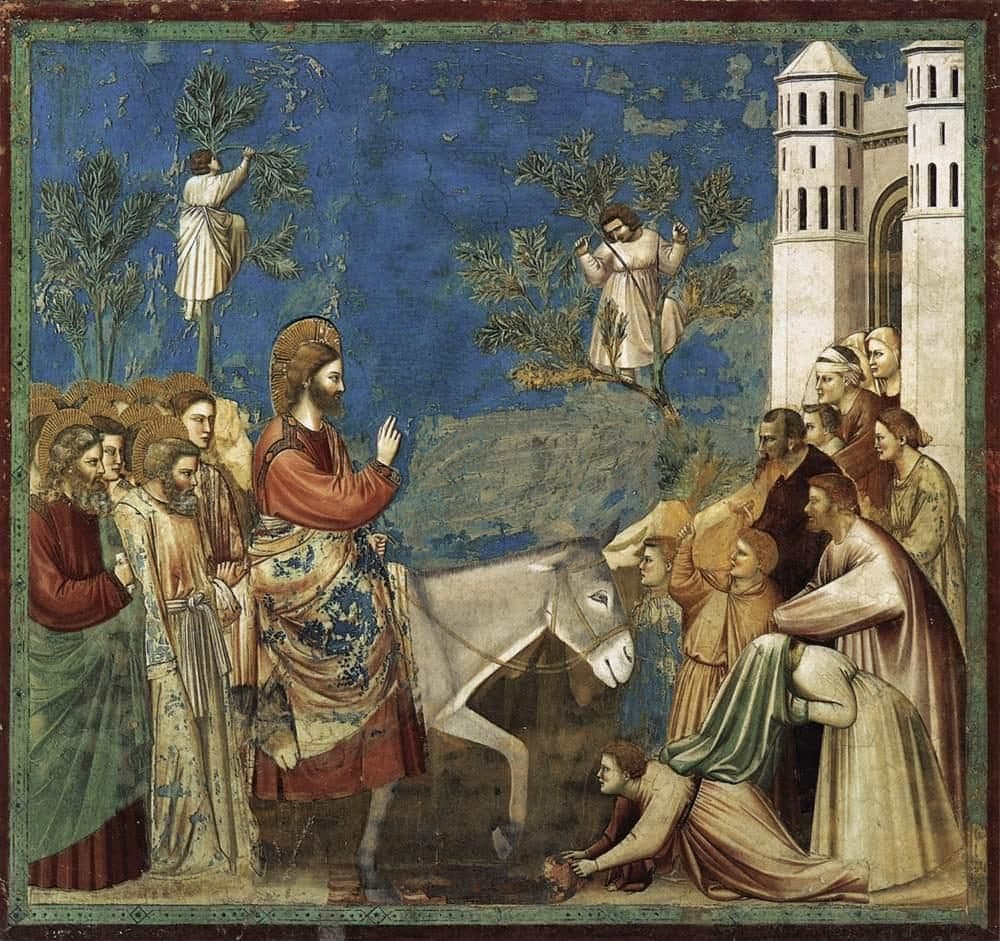 Jesus rides a donkey toward a city gate with his hand raised in blessing; the apostles follow, two men cut branches in the trees, and people at the gate lay their cloaks before the donkey.