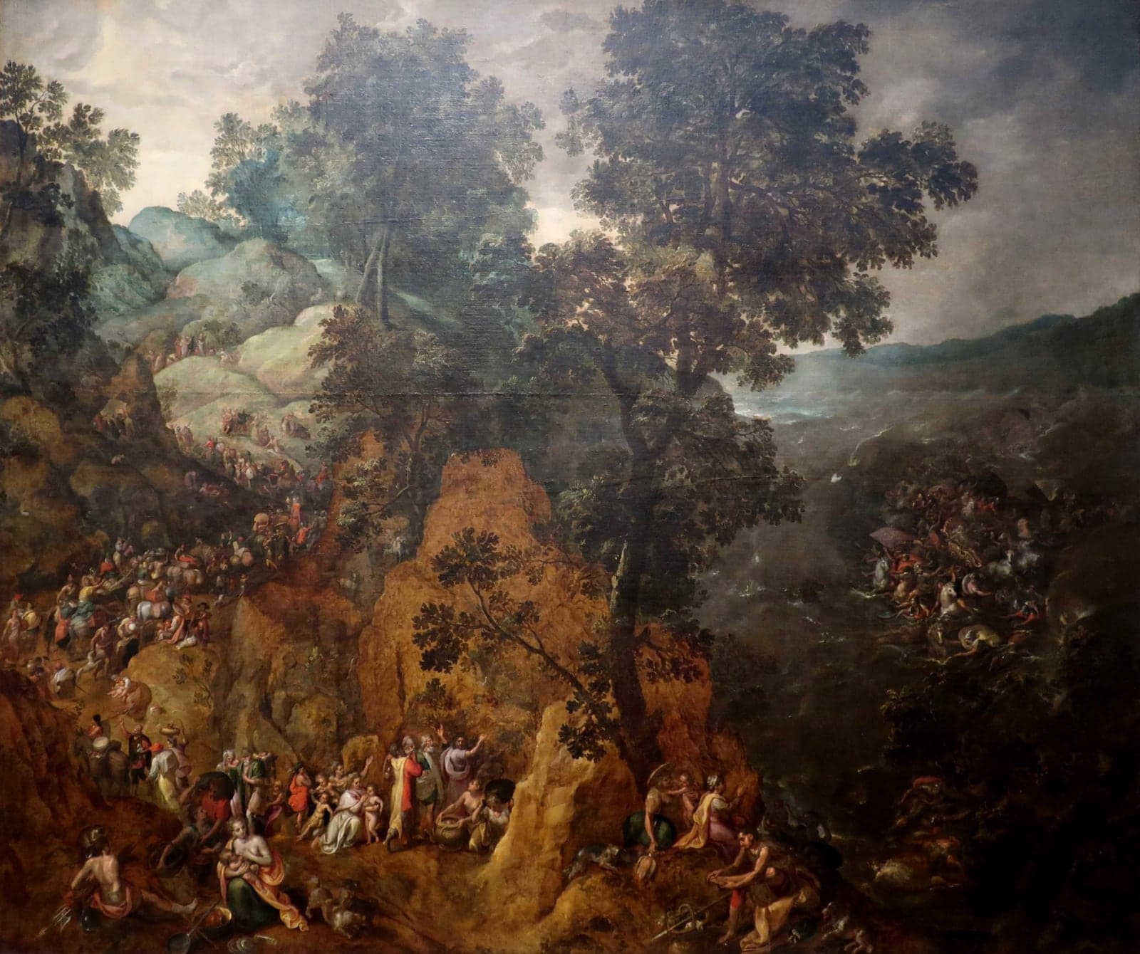 A wooded mountain landscape under a stormy sky, with a long column of people, riders and pack animals winding up a rocky road on the left, families resting among the rocks in the foreground, and horsemen struggling in rough water on the right.