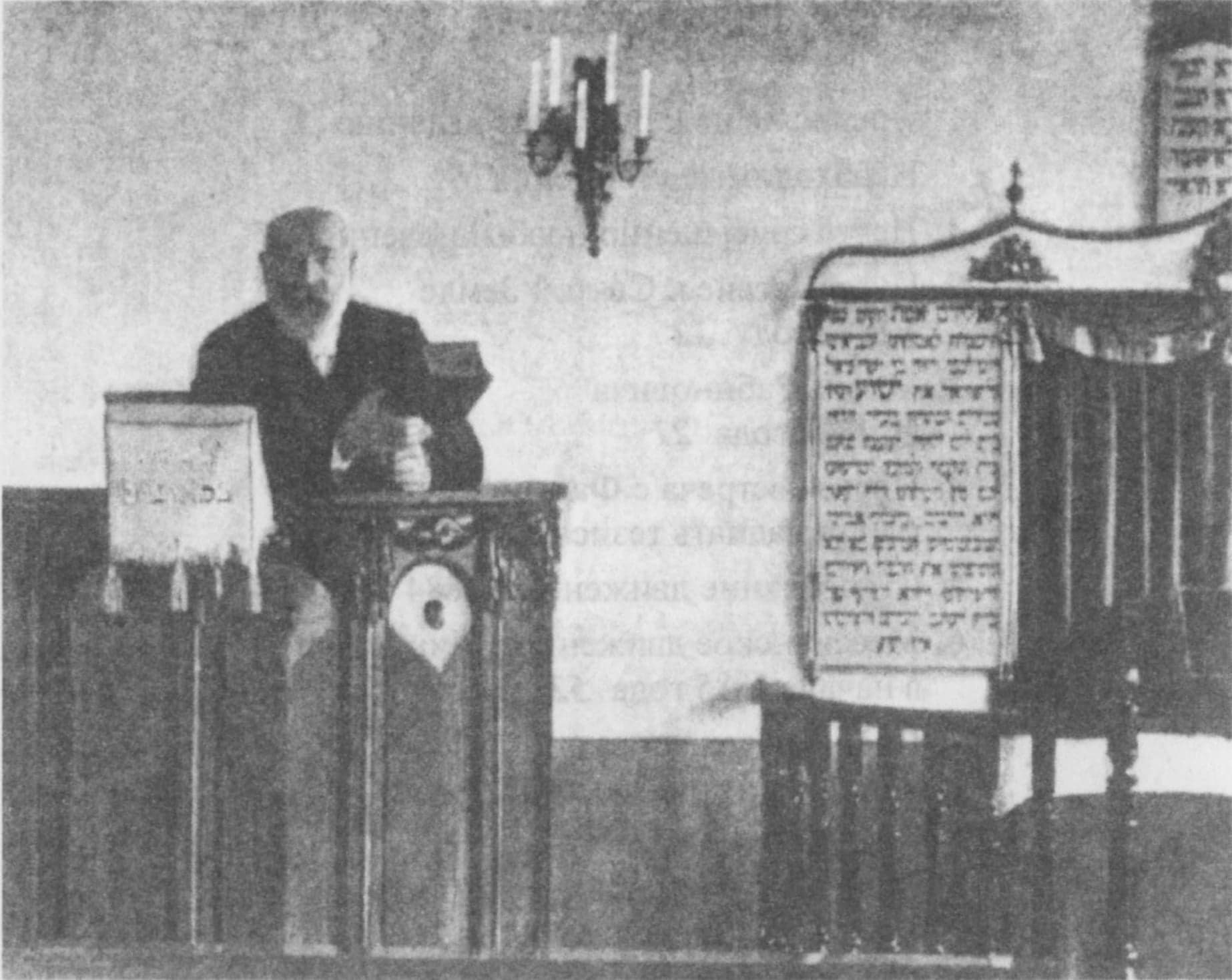 A grey printed picture of a bearded man in a dark coat standing at a lectern in a hall, with tablets bearing Hebrew letters on the wall behind him.