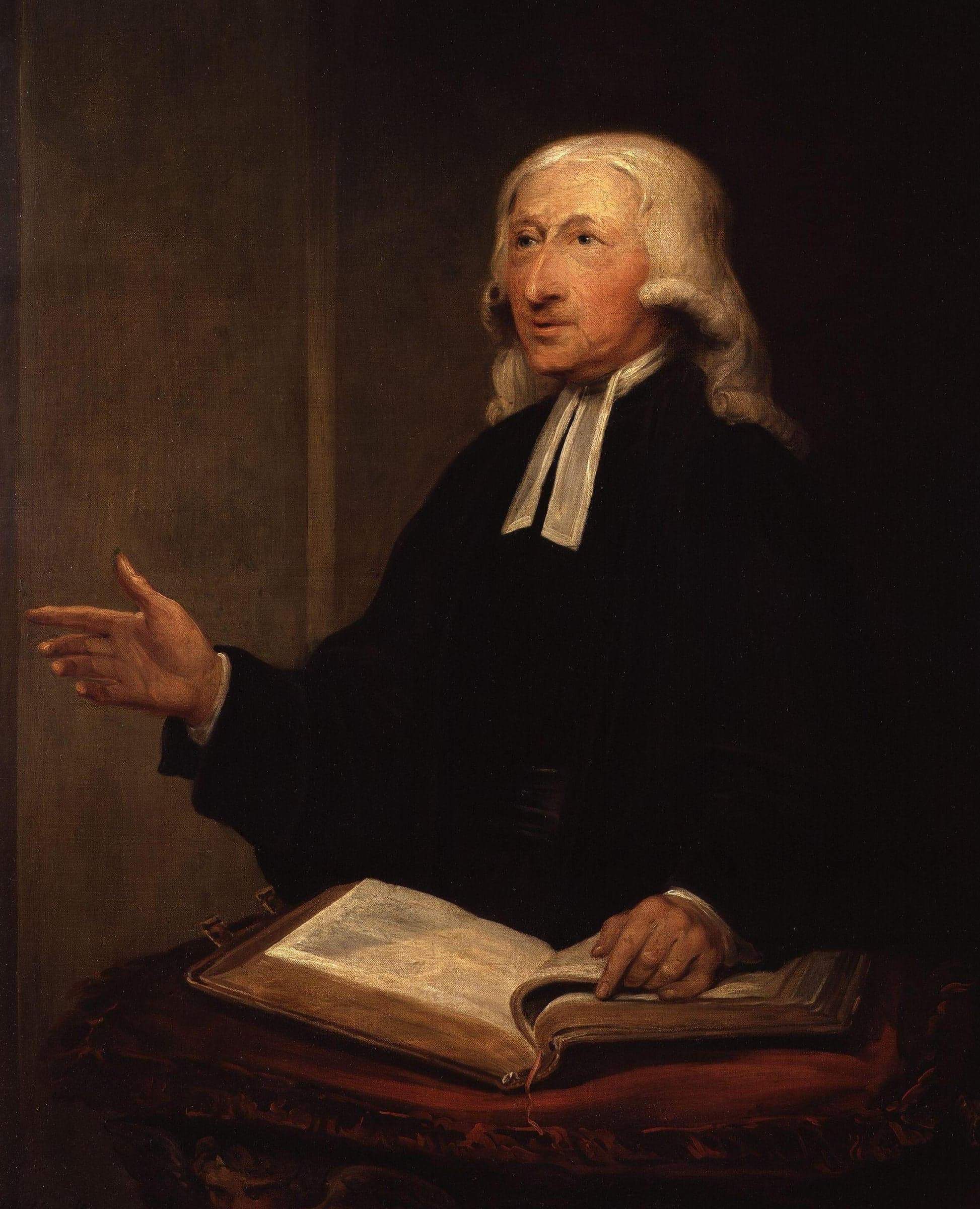 John Wesley in old age, in a black gown and white bands, with long white hair, raises his right hand as he speaks over an open Bible on a red cushion.