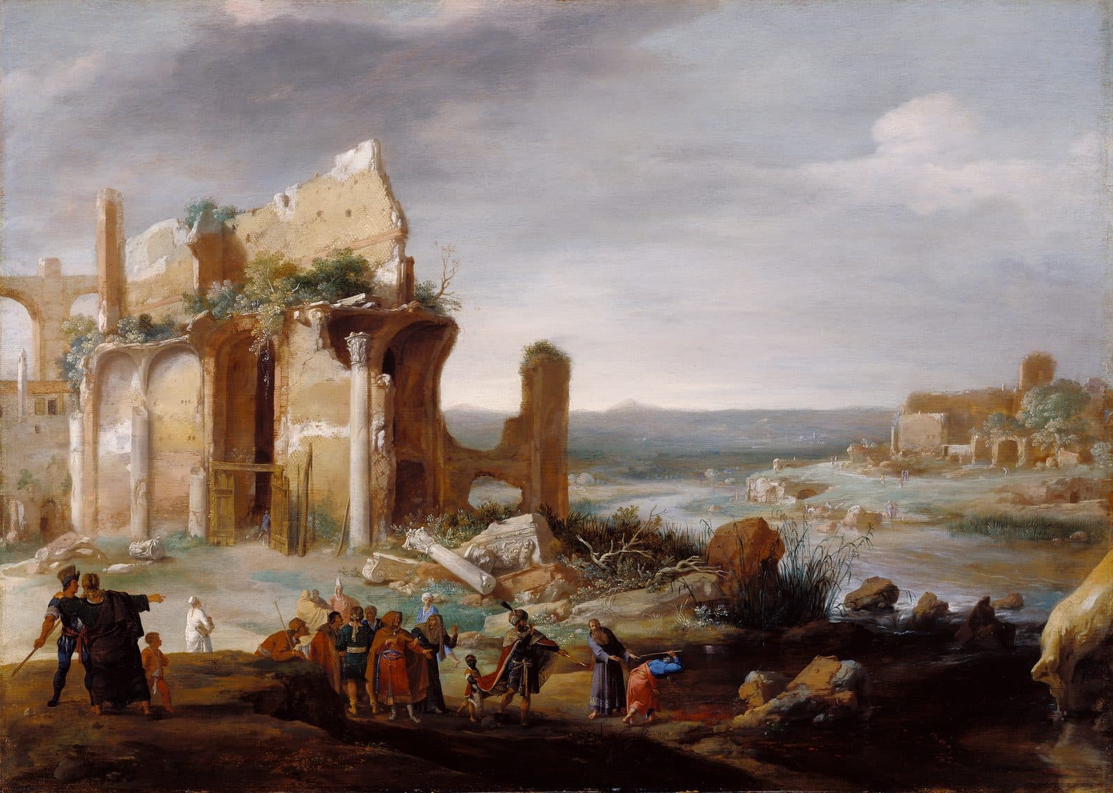 Beside tall ruins on a riverbank, a robed man bends to touch the water with a staff while a crowd in bright robes and turbans watches, and the river runs off into a wide valley.