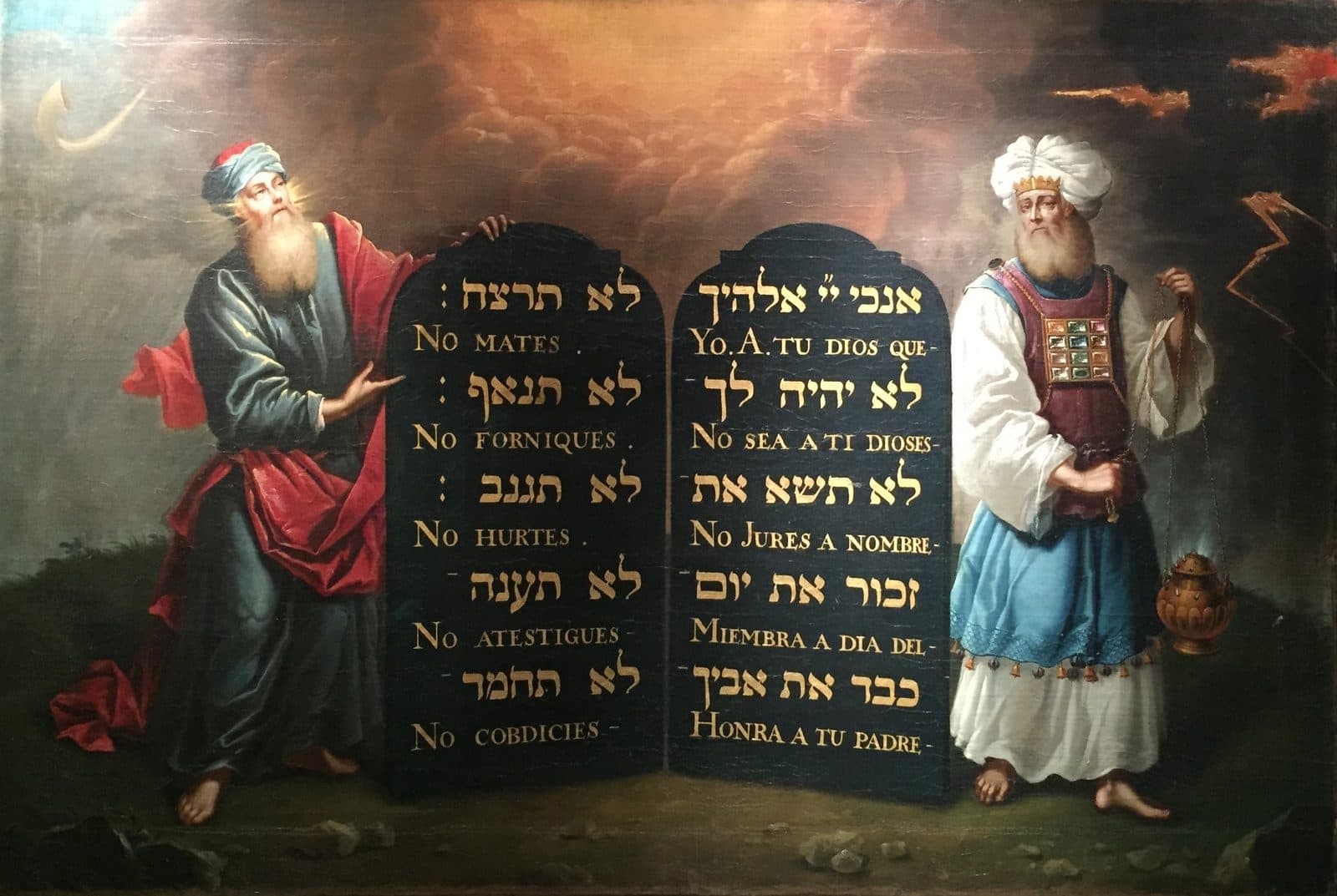 Moses in a red cloak at left and Aaron in a white turban and a jeweled breastpiece at right stand beside two round-topped black tablets. Each tablet carries five lines of gold Hebrew with a line of Spanish under each, below a stormy sky with lightning.
