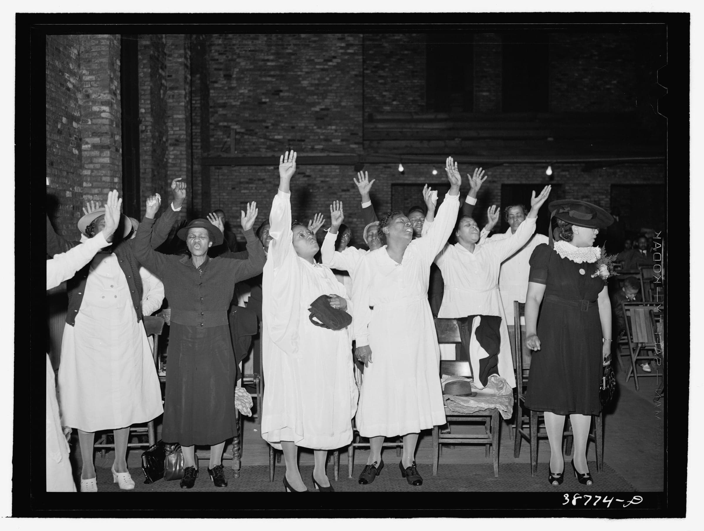 Women of a Pentecostal church in Chicago stand among wooden chairs with their hands raised, singing, under a brick wall.