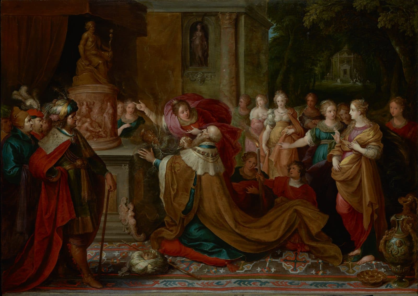 Solomon kneels in a gold robe before an idol on a pillar while his wives stand around him and one points him to it