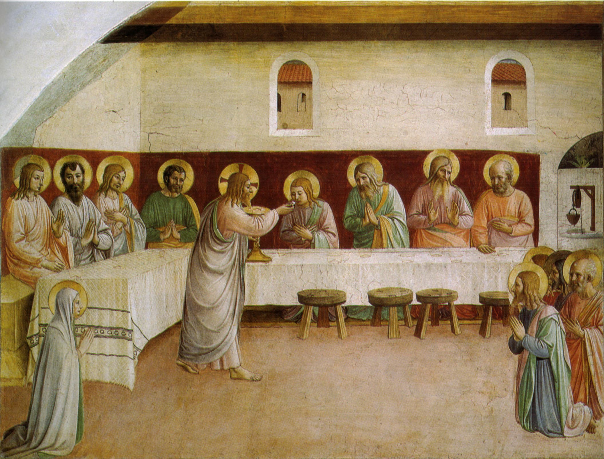 A fresco of a long white table in a plain room with two small windows. Christ, standing and barefoot, holds a chalice and places bread in the mouth of an apostle seated at the table, while seven more haloed apostles sit behind it with hands joined in prayer. On the right four apostles kneel together, and on the left a woman in a grey mantle kneels with her hands raised.