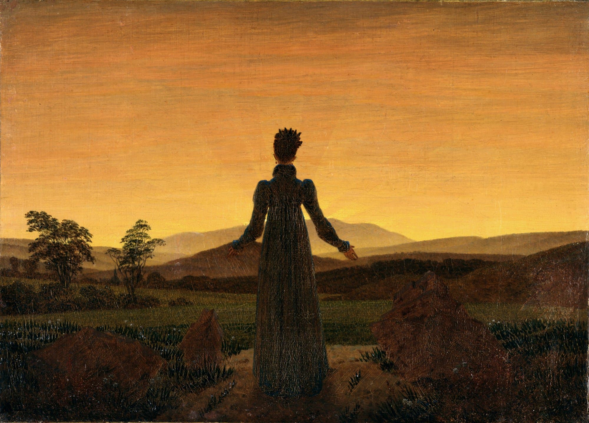 A woman in a long dark dress stands with her back to the viewer on a path between rocks, her arms open toward a low sun over far hills under a golden sky.