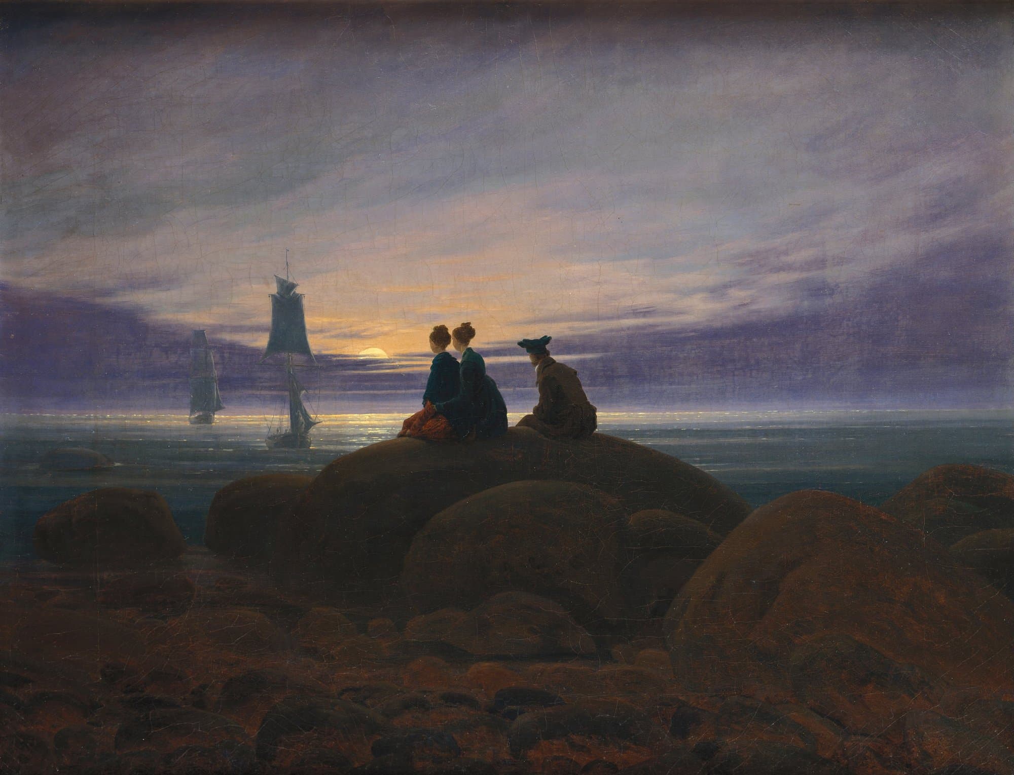 Two women and a man in a cap sit with their backs turned on a large rounded boulder above a calm sea at dusk, watching the moon rise on the horizon under a purple and gold sky, while two sailing ships come in toward the shore and dark rocks fill the foreground.
