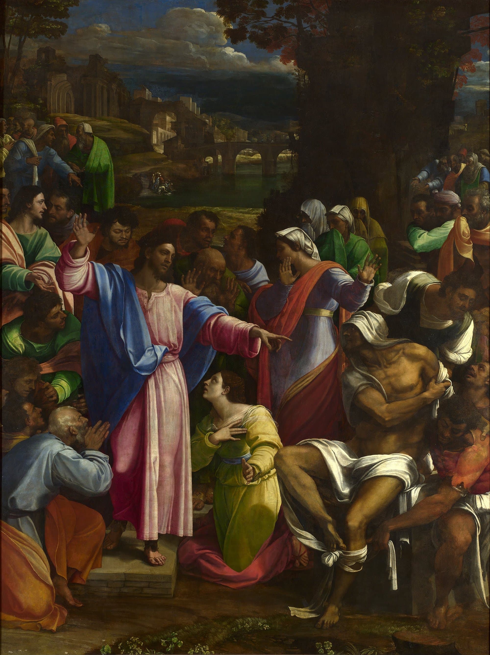 A crowded scene before a town and a bridge under a stormy sky: Jesus in a pink robe and blue mantle raises one hand and points with the other at Lazarus, a gaunt man seated at the right pulling off his grave cloths, while a woman in yellow kneels at Jesus' feet and a woman in red raises her hands, and onlookers press in on every side.