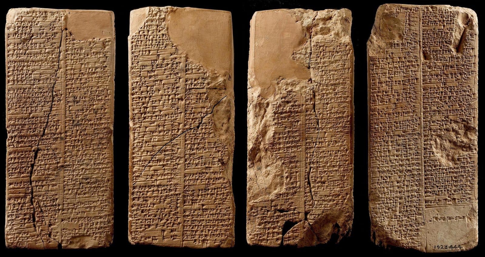 Four faces of a tall clay prism photographed side by side, each covered top to bottom in columns of wedge-shaped cuneiform signs, with cracks and broken patches in the clay
