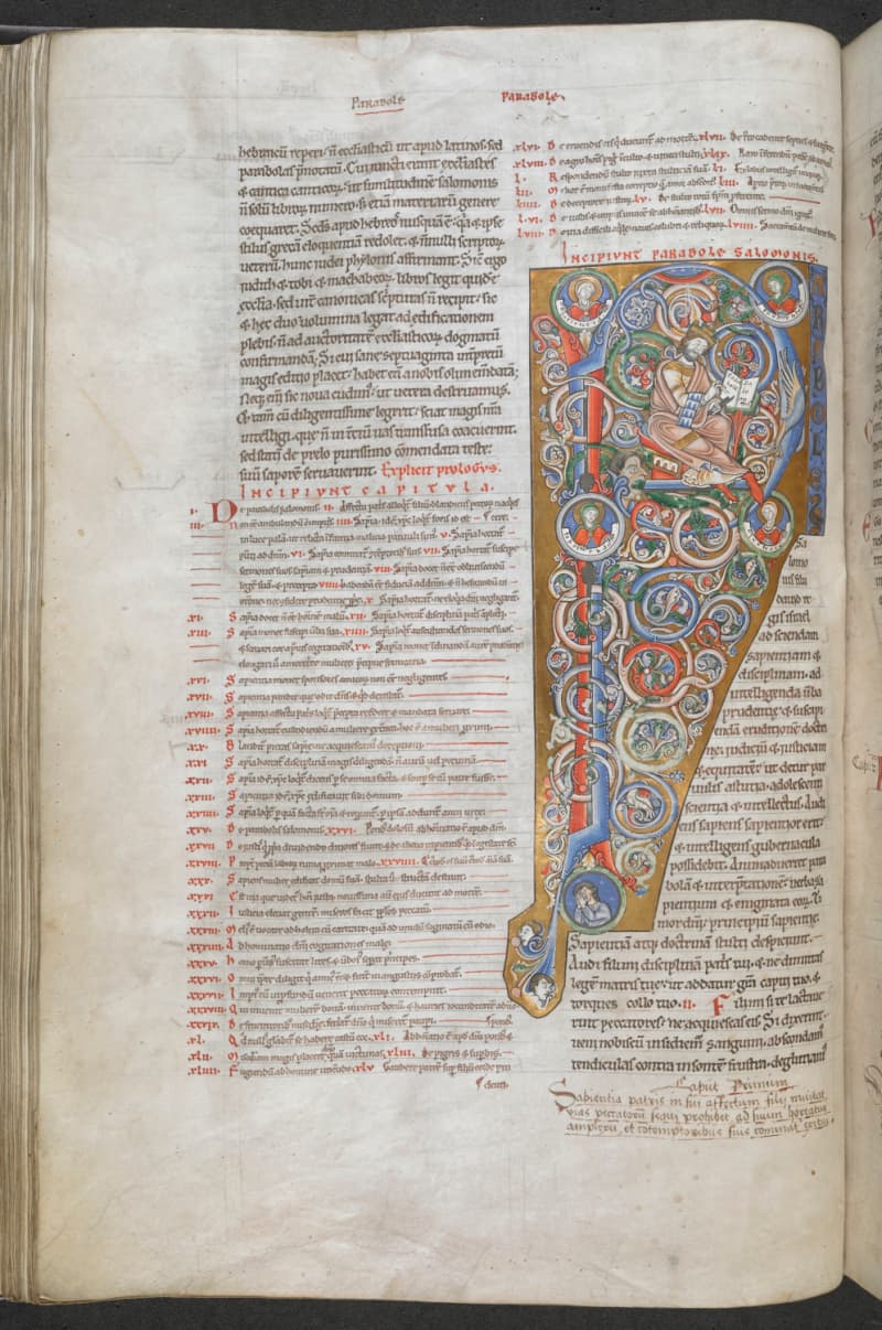 A page of a twelfth-century Latin Bible in two columns of script, with a tall initial letter of blue, red and gold scrolls in which a crowned king sits writing in a book, ringed by small round portraits.
