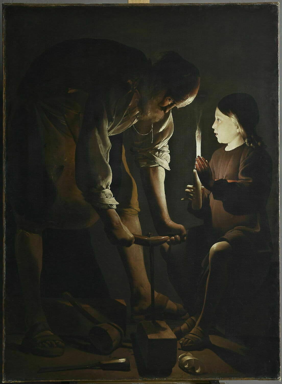 In a dark workshop an old man bends over a beam of wood, driving an auger into it with both hands. A young boy sits beside him holding up a candle, its light falling on the man's face and arms.