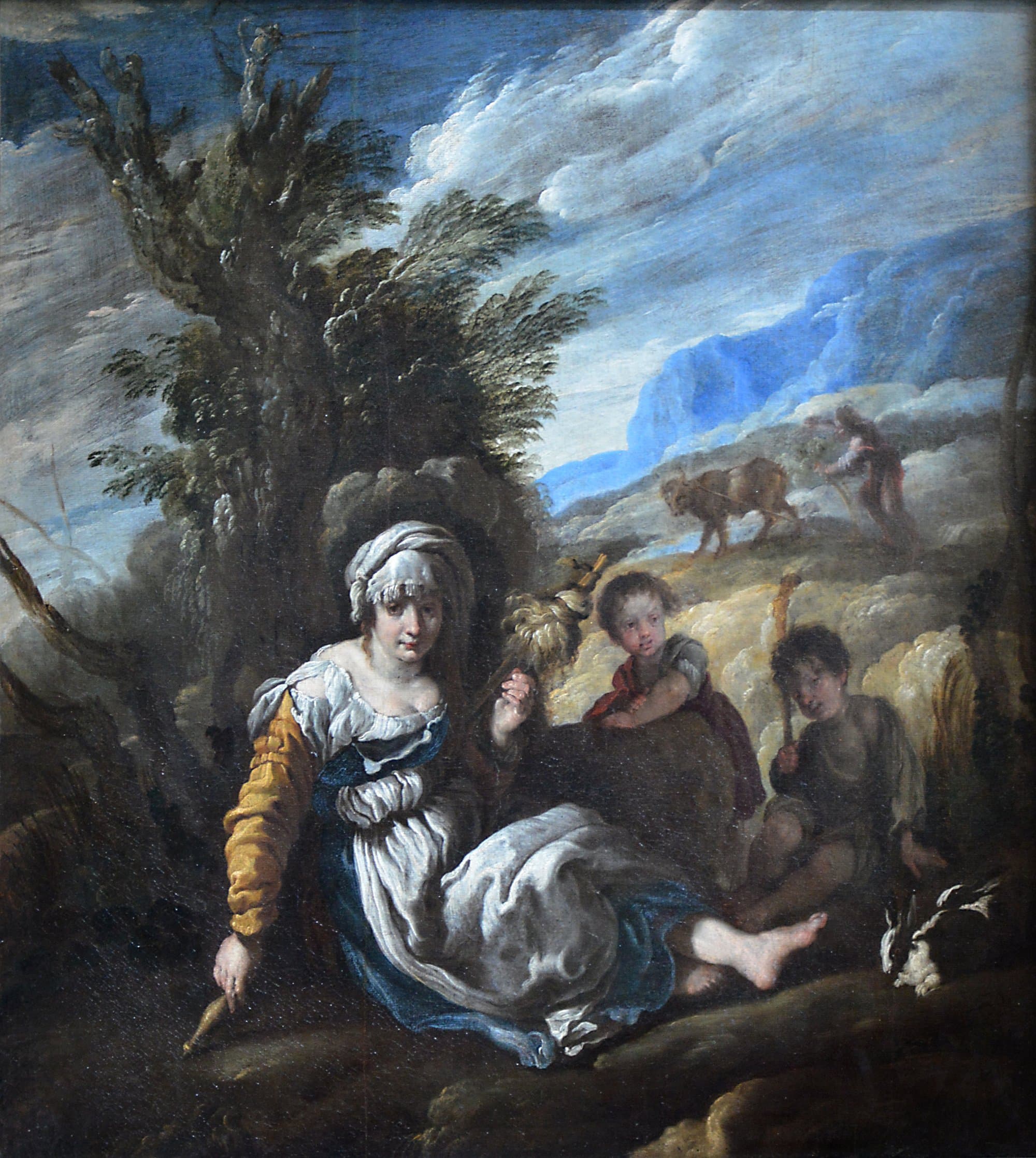 A young woman in a white headscarf sits on the ground under a tree with a distaff of wool and a spindle, two small boys sit beside her with a rabbit at their feet, and on the hillside behind them a man drives a plough drawn by an ox.