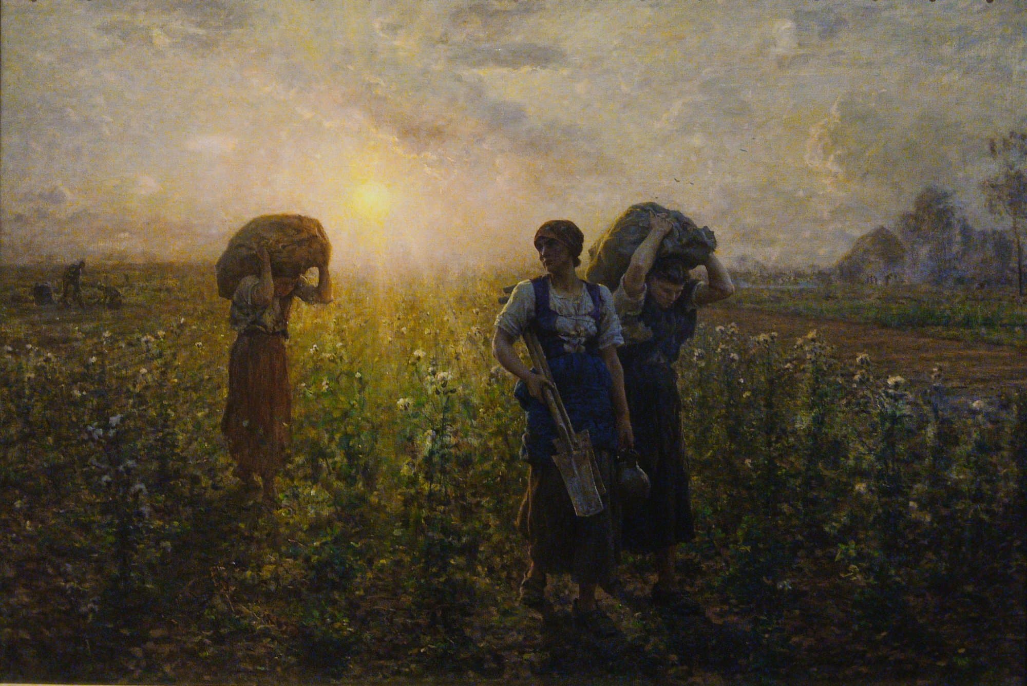 Three women walk home through a field of thistles at sunset, two of them bent under heavy sacks carried on their heads and one with a spade and a jug in her hands, while the low sun glows behind them.