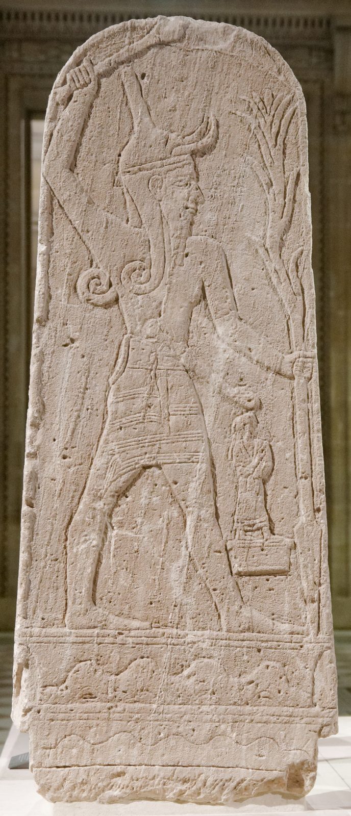 A round-topped limestone stele in low relief: a striding god in a horned crown raises a club over his head and holds a branching spear, with a small robed figure on a pedestal before him.