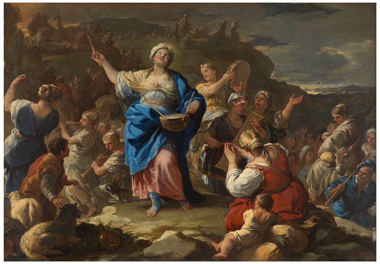 A woman in a white turban, a blue mantle and a pink skirt beats a drum hung at her waist and looks up to the sky. Women around her play a tambourine, flutes and a harp, children and sheep sit on the rocks at her feet, and a line of people with camels crosses the hill behind under a stormy sky over the sea.